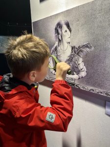 Ein kleiner Gast der Fine-Art Ausstellung in Buchholz-Sprötze schaut sich mit einer Lupe ein gemaltes Frauen Portrait von Sara Heinrich an. Die Künstlerin stellte ihre werke im Rahmen der Heidekultour 2021 aus.